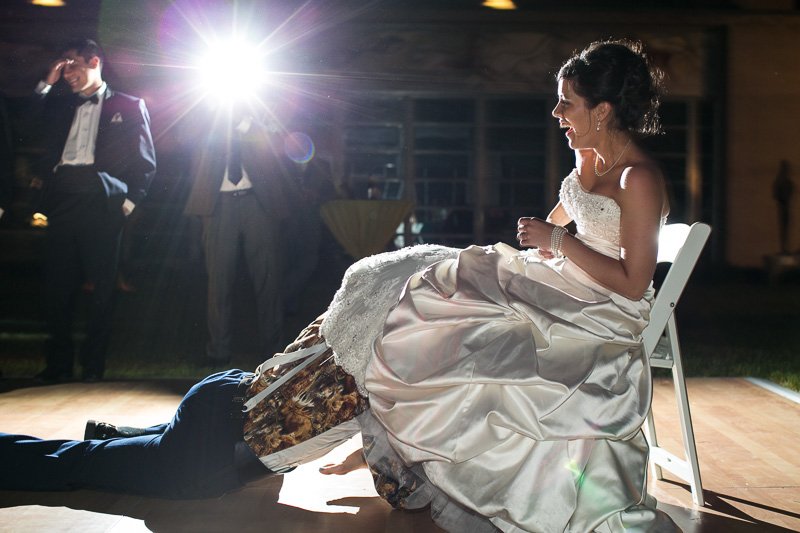 Bride sitting on a chair during her wedding reception, smiling, with a person lying on the floor in front of her. Two men in tuxedos are standing in the background, and a bright light source creates a lens flare.