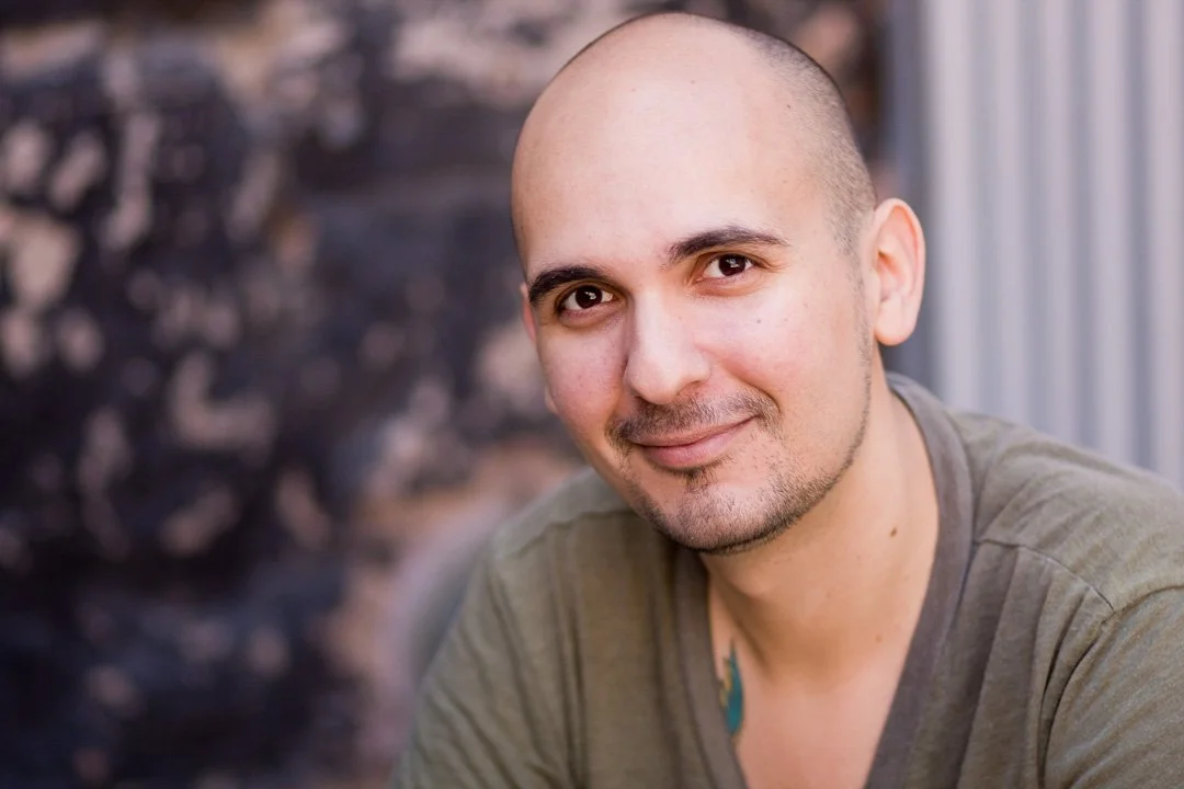 Close-up of a smiling, bald man with a light beard, wearing a green T-shirt, outside near a metallic fence.