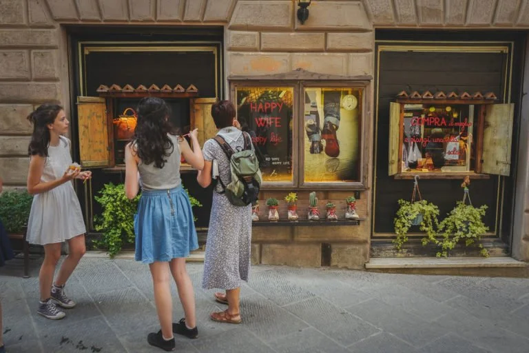 Three women standing on a sidewalk in front of a storefront with potted plants and window displays.