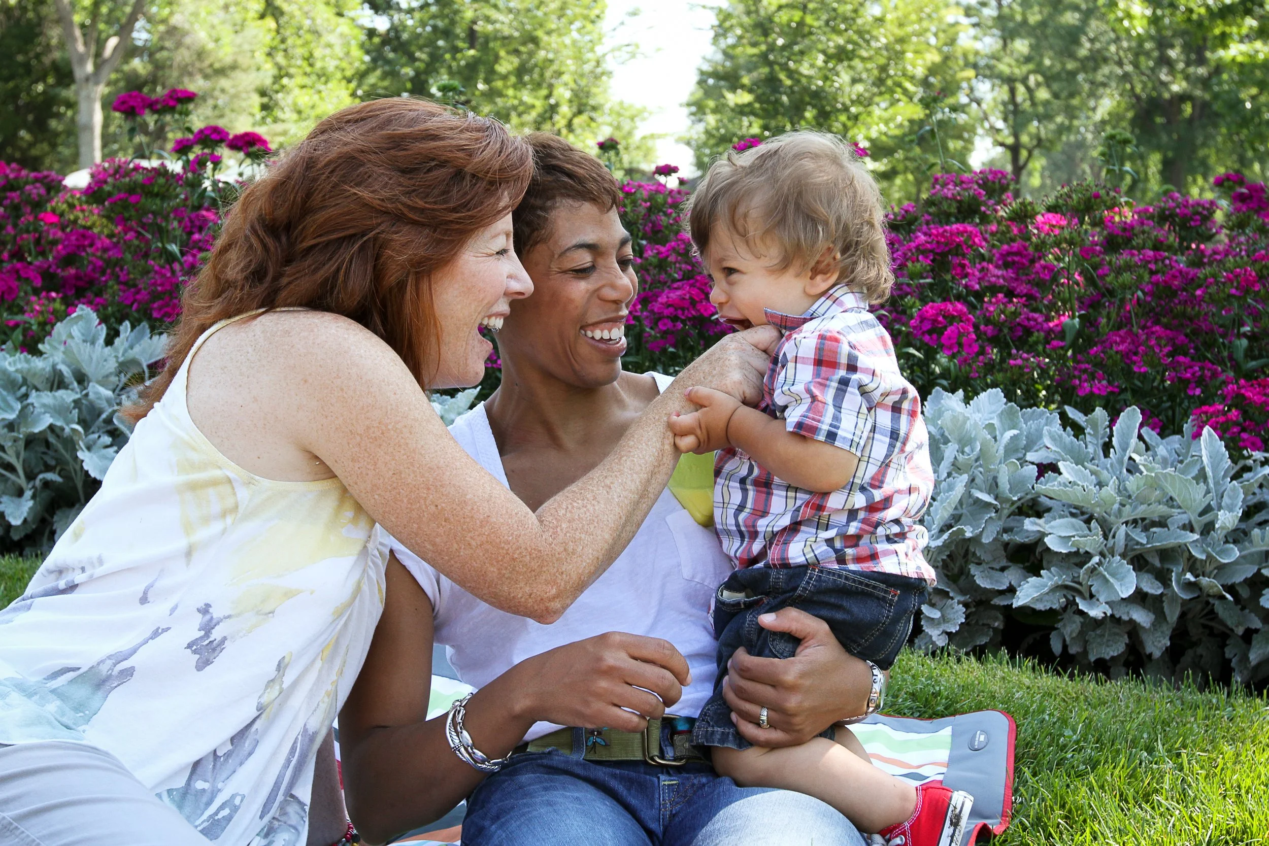 Three people, two women and a young boy, laughing and playing together outdoors in a garden with vibrant pink and purple flowers and green trees in the background.