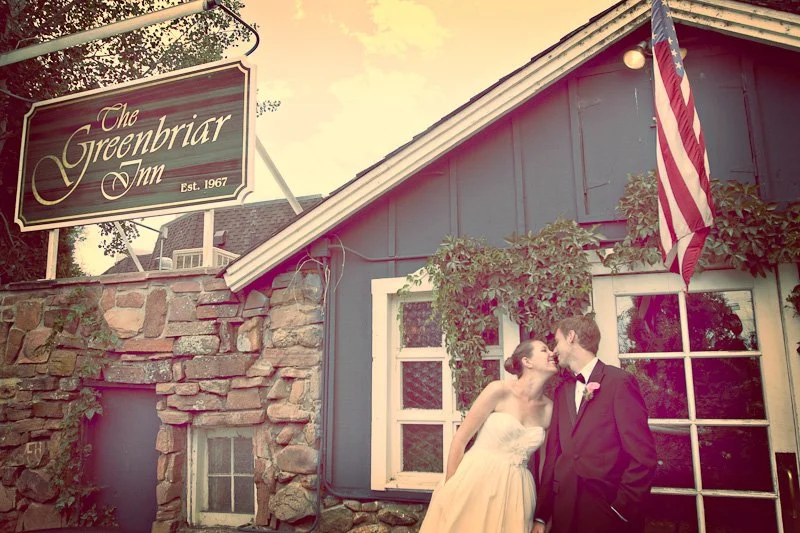A newlywed couple leaning in for a kiss in front of the Greenbriar Inn, a stone and wood-constructed building with an American flag and greenery, during sunset.