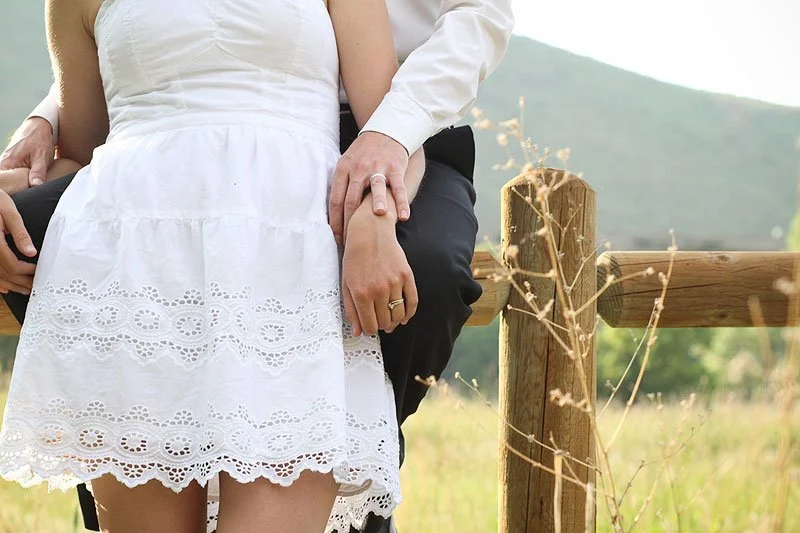A close-up of a couple sitting outdoors on a wooden fence, with a mountain and greenery in the background. The woman is wearing a white dress with lace details, and the man is wearing a white shirt and black pants. The woman is wearing a ring on her 