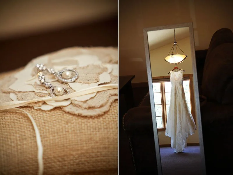 Close-up of pearl earrings with rhinestones on a textured surface and a wedding gown hanging in a mirror reflection.