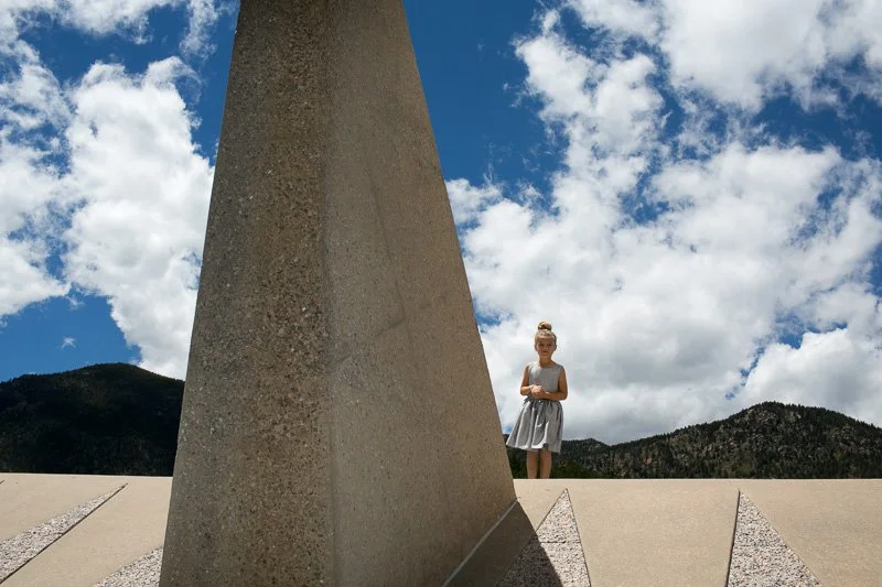 A person standing outdoors, with a large concrete or stone structure in the foreground, mountains in the background, and a partly cloudy sky.