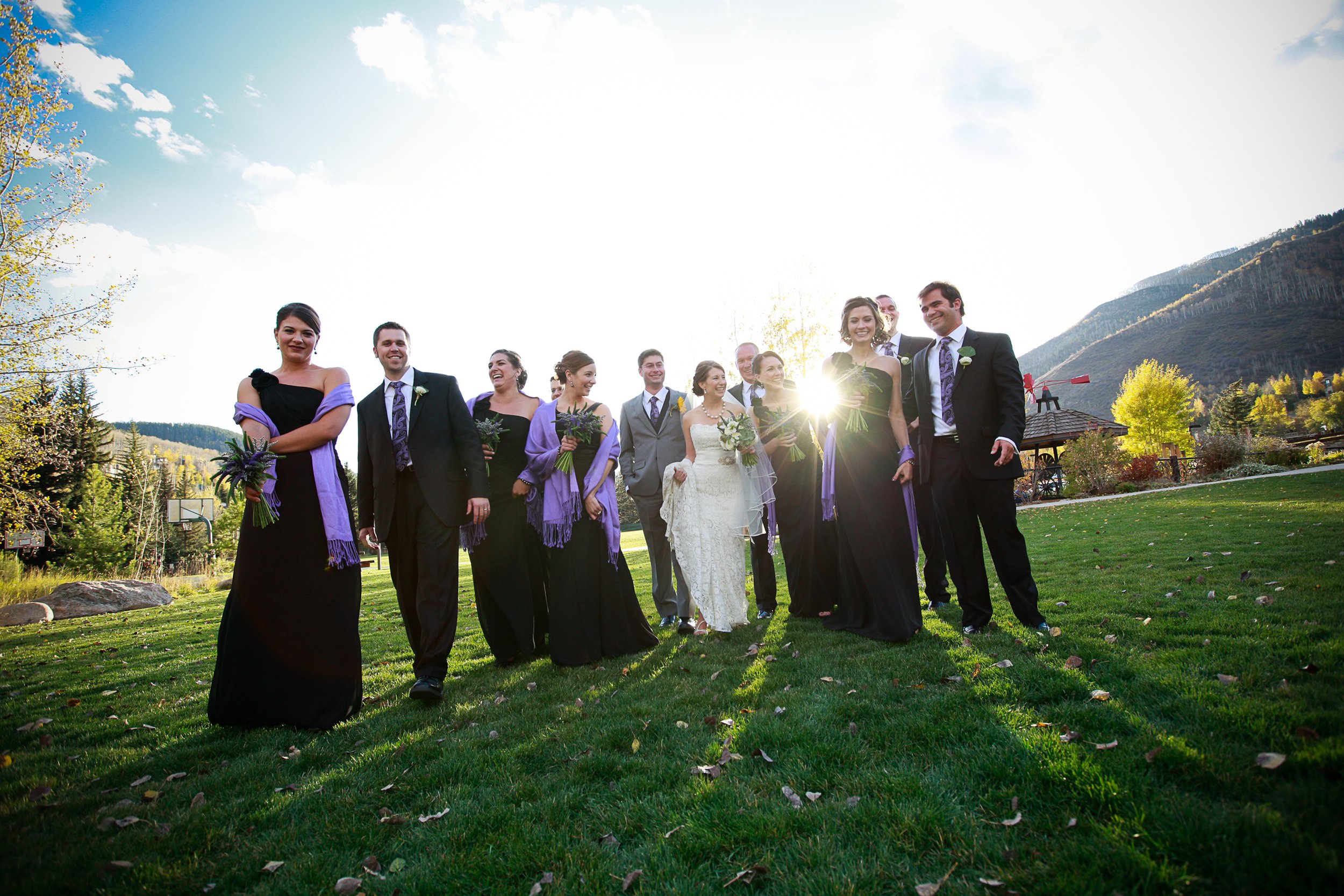 A wedding party of eleven people, including the bride in a white dress holding a bouquet and ten bridesmaids and groomsmen, walking on green grass outdoors with mountains in the background. The sun is shining, creating a bright glow.