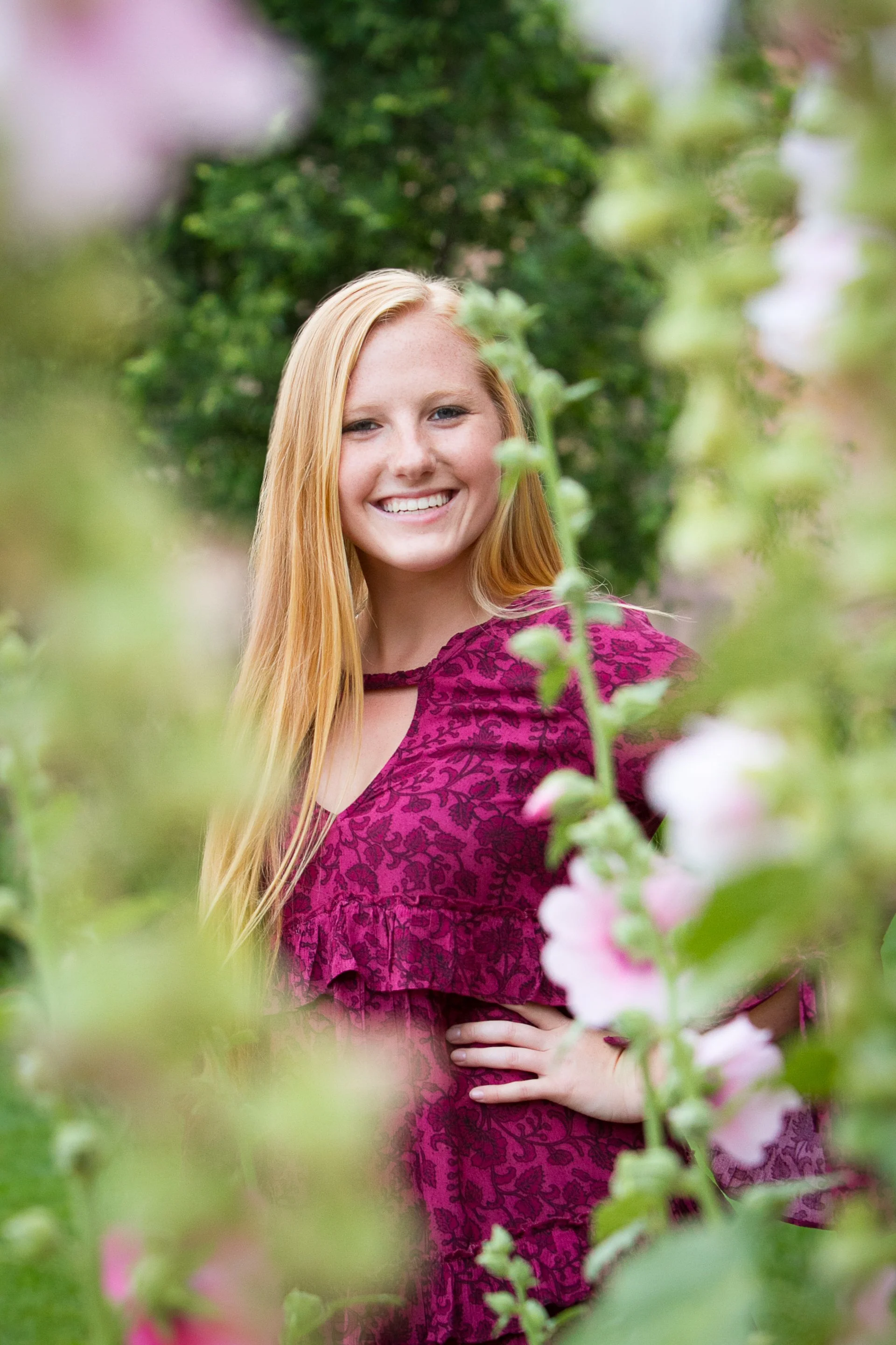 A young woman with long blonde hair, wearing a purple patterned dress, smiling and standing in a lush garden with green foliage and pink flowers.