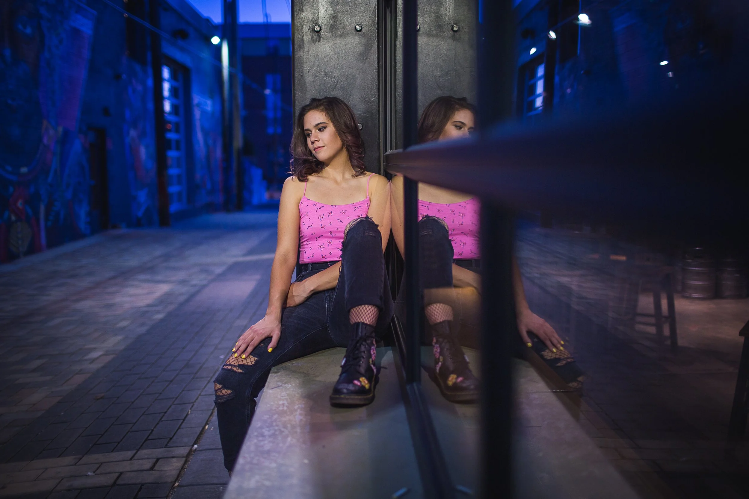 A young woman with brown hair wearing a pink top and ripped jeans, sitting on a bench next to a reflective glass wall in a dimly lit urban alley with graffiti.
