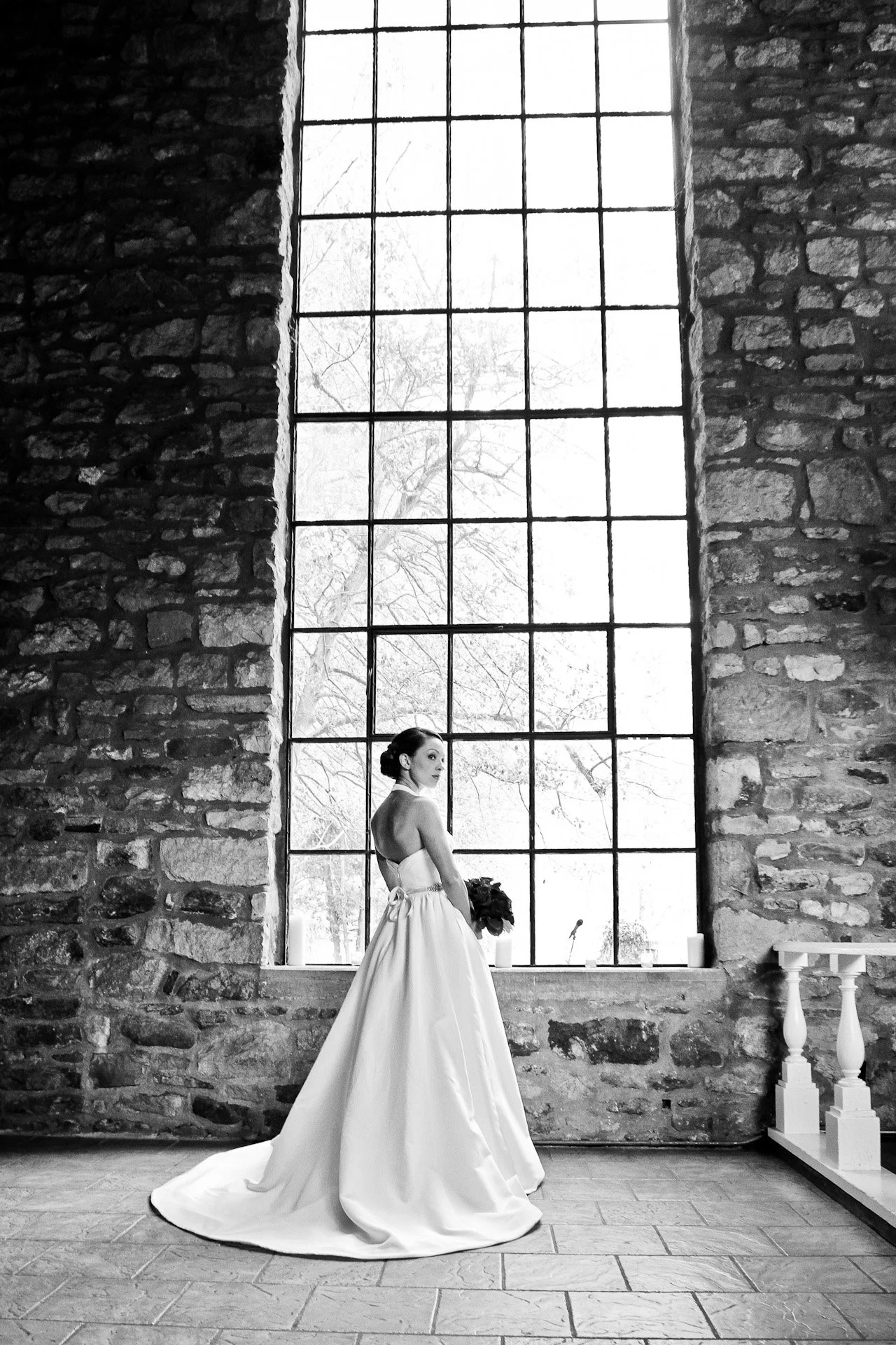 Bride in wedding dress holding bouquet stands inside a rustic stone building by a large grid window with bare trees visible outside.