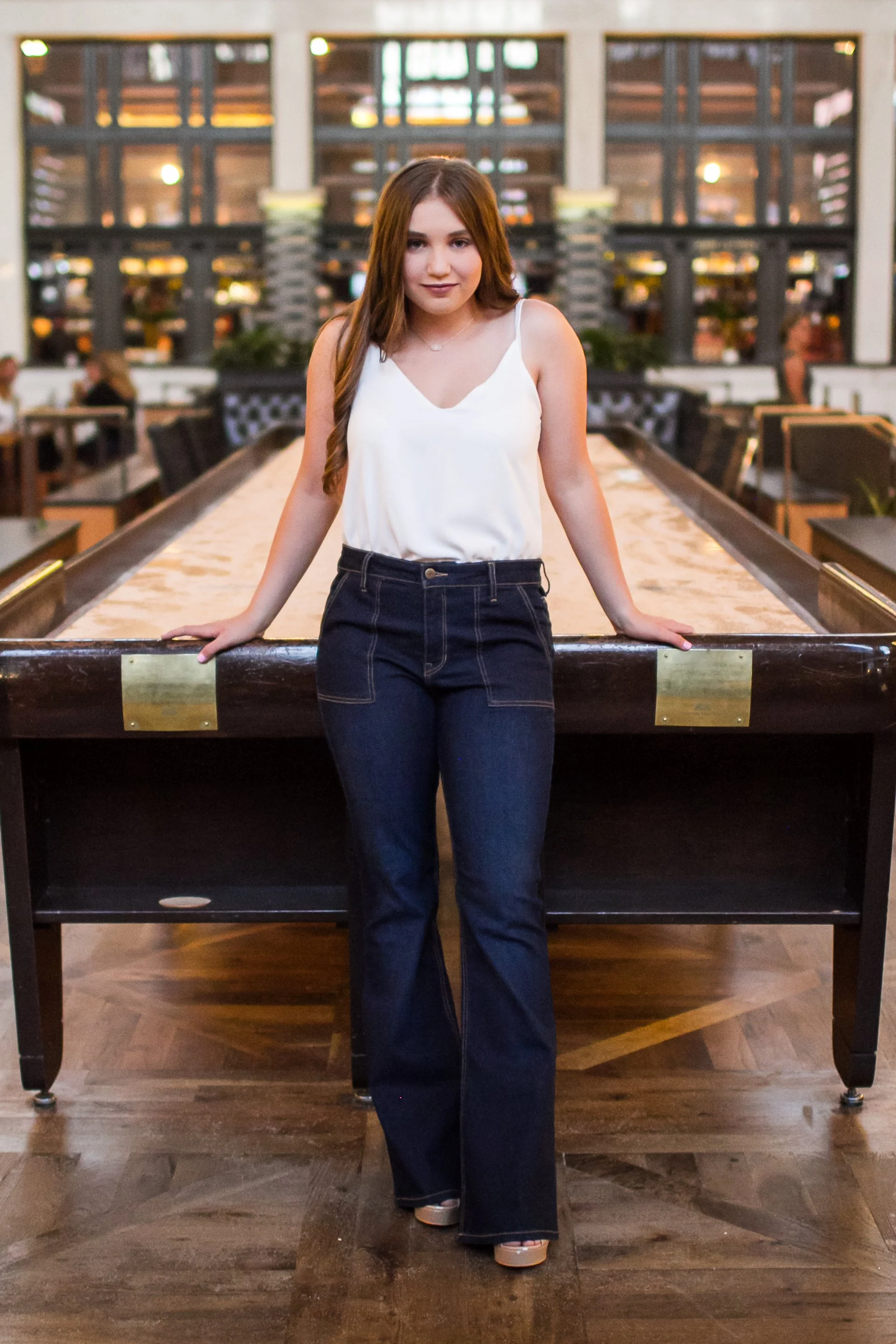 A young woman with long, wavy brown hair in a sleeveless white top and high-waisted dark jeans stands behind a shuffleboard table in a spacious indoor recreational area with large windows and seating in the background.