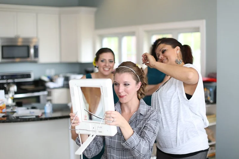Three women in a kitchen, with one sitting and holding a mirror, while the others stand behind her, smiling and hairstyling her hair.