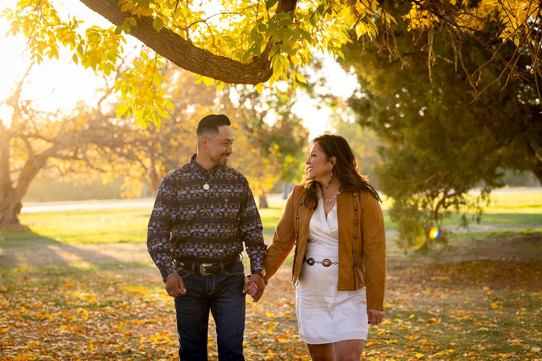 A couple holding hands, walking through a park with autumn leaves on the ground, trees with yellow and green leaves, and sunlight filtering through the branches.