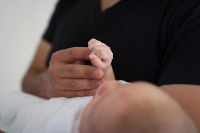 A person with hands clasped together resting on another person's arm, who appears to be a baby or young child.