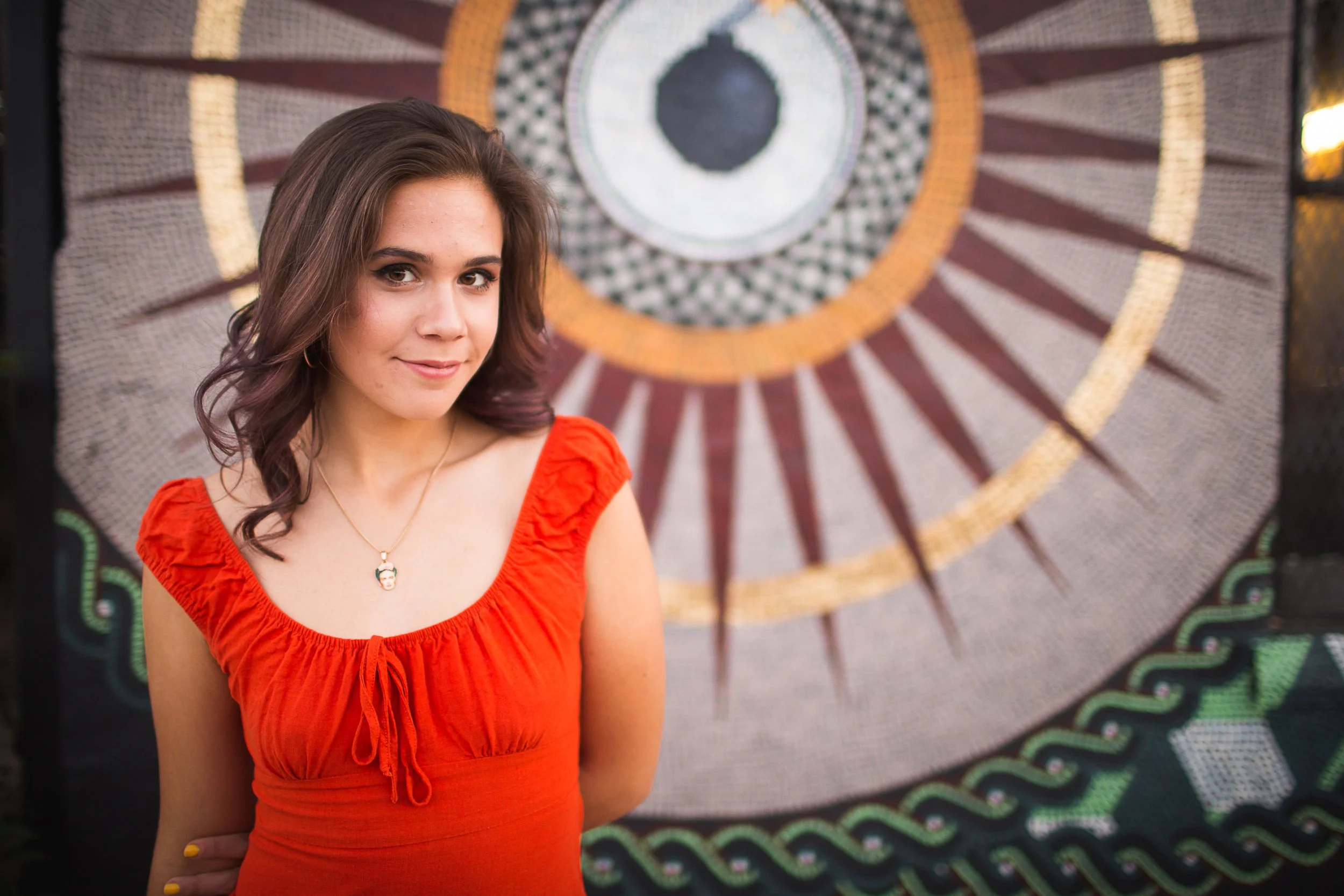 A young woman with brown hair wearing a red sleeveless top and a gold necklace standing in front of a decorative circular mosaic wall with concentric patterns and geometric designs.