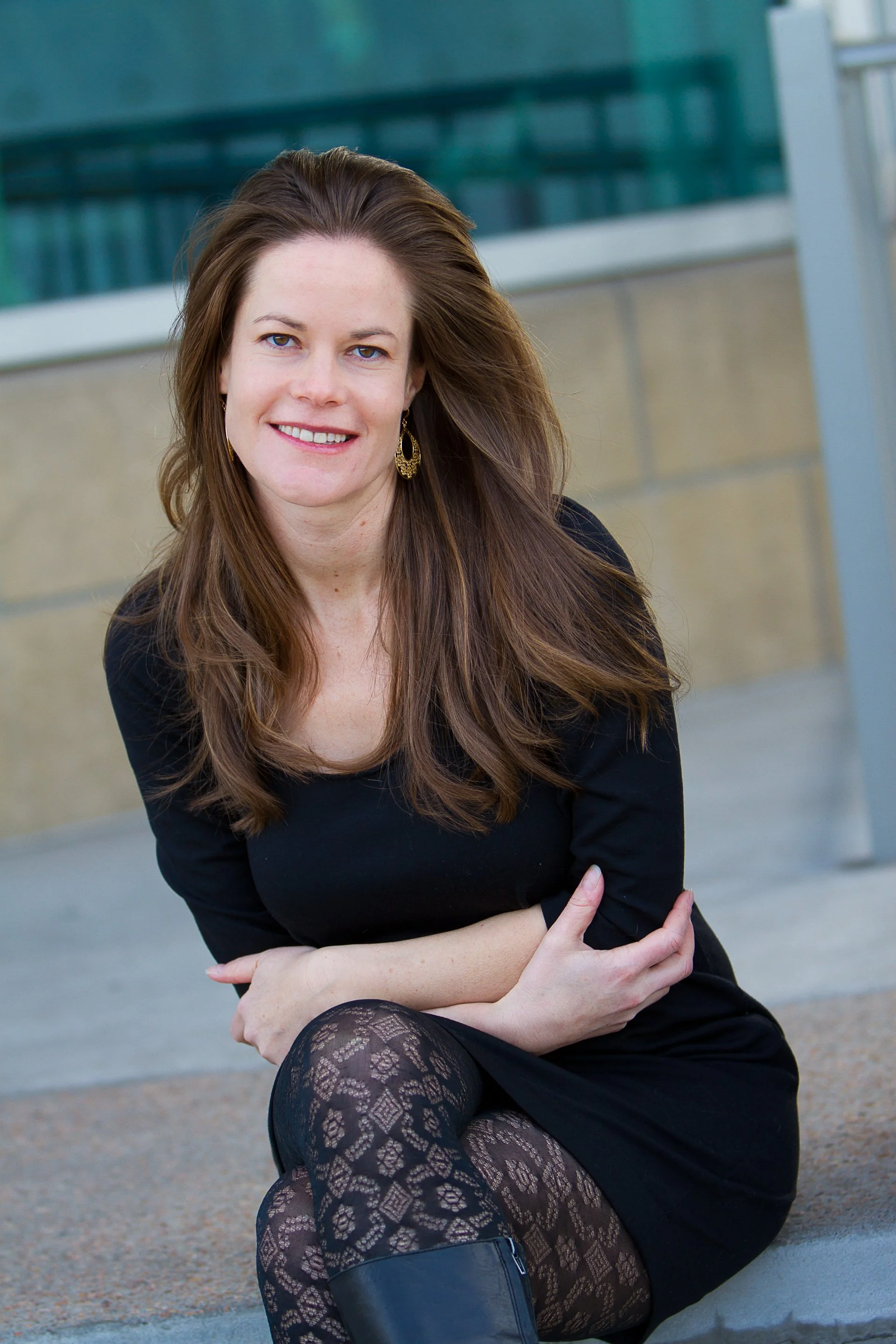 A smiling woman with long brown hair, wearing a black dress, patterned tights, and boots, sitting outdoors in front of a modern building.