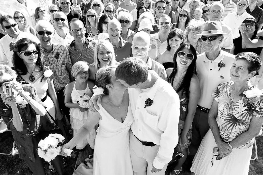 A large crowd of people gathered outdoors, celebrating a wedding with the bride and groom sharing a kiss in the center.