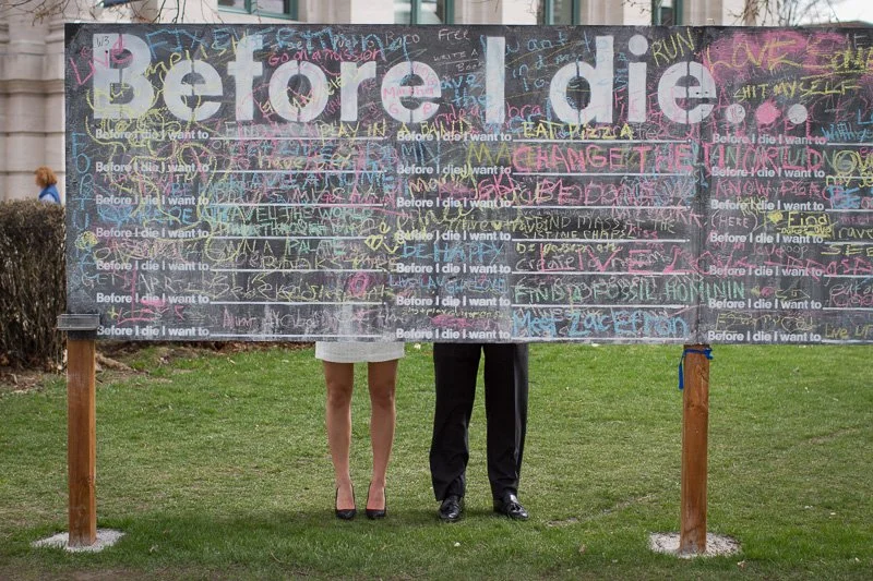 A large chalkboard with the words "Before I die" written in large letters, filled with colorful handwritten responses from various people. The chalkboard is outdoors, with legs supported by wooden posts. People are standing behind it, with only their