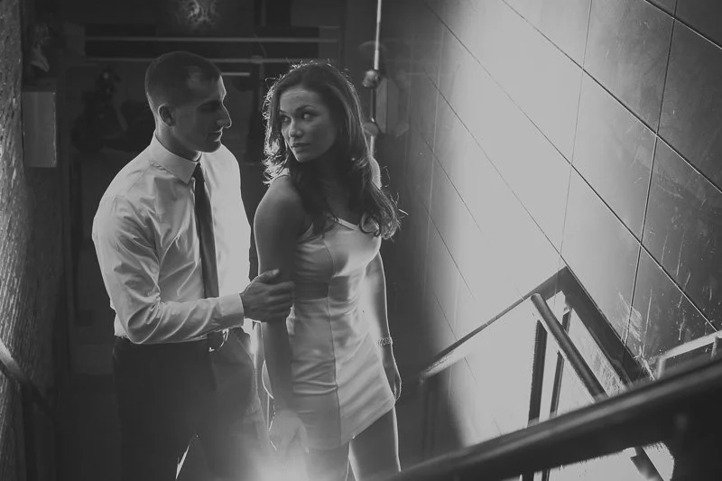 A man and woman standing on a staircase in a dimly lit setting, with the man gently holding the woman's arm and both looking at each other.