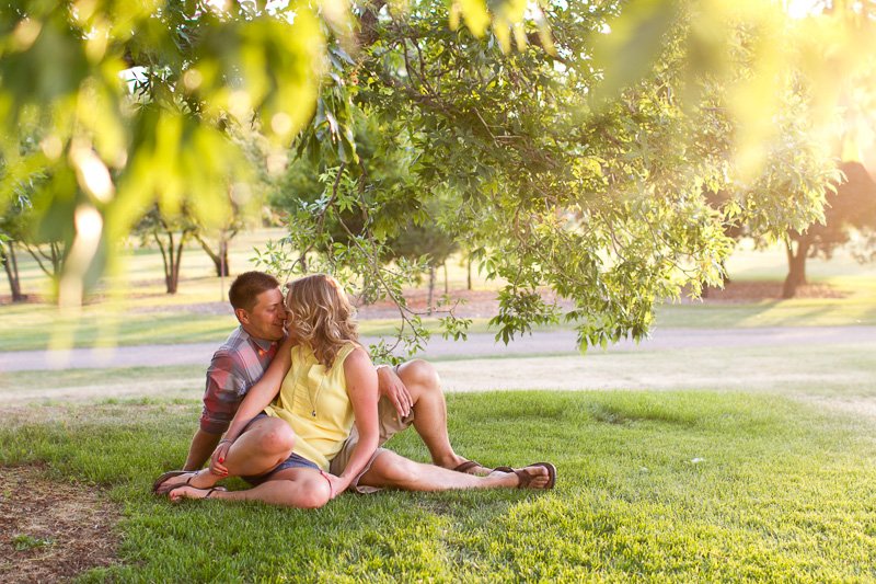 A couple sitting on the grass under a leafy tree, leaning in for a kiss in a park during late afternoon