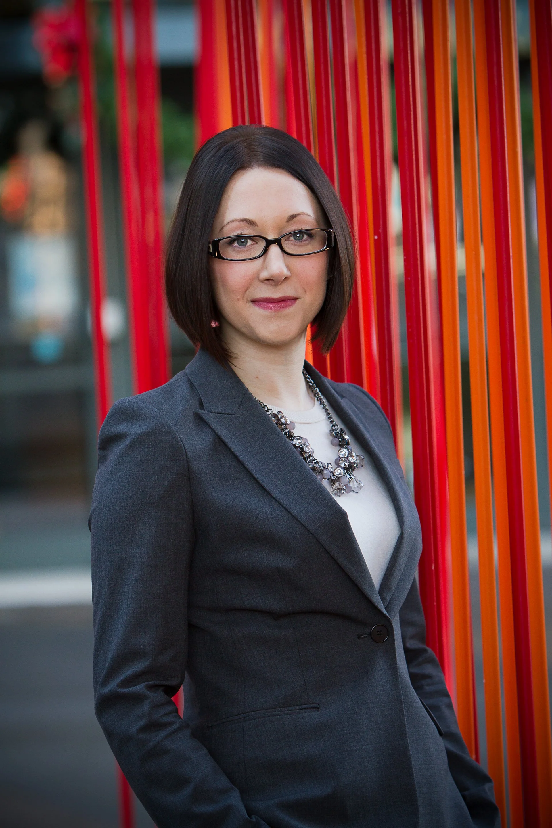 A woman with shoulder-length dark hair wearing glasses, a gray blazer, and a statement necklace stands outdoors against a background of red vertical bars.