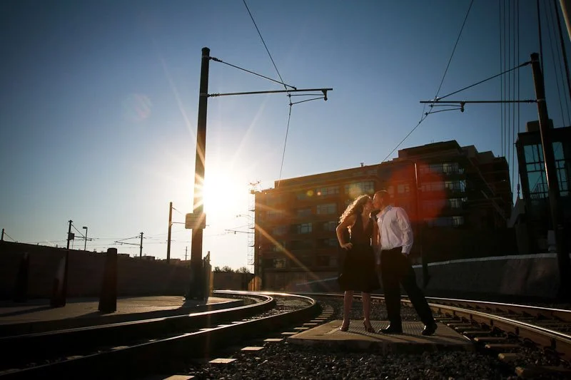 A couple standing on train tracks near overhead electrical lines with a building in the background during sunset.