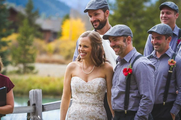 A bride in a white lace dress smiling with a group of men dressed in gray shirts and suspenders, all wearing gray caps, outdoors in a scenic setting with trees and mountains in the background.