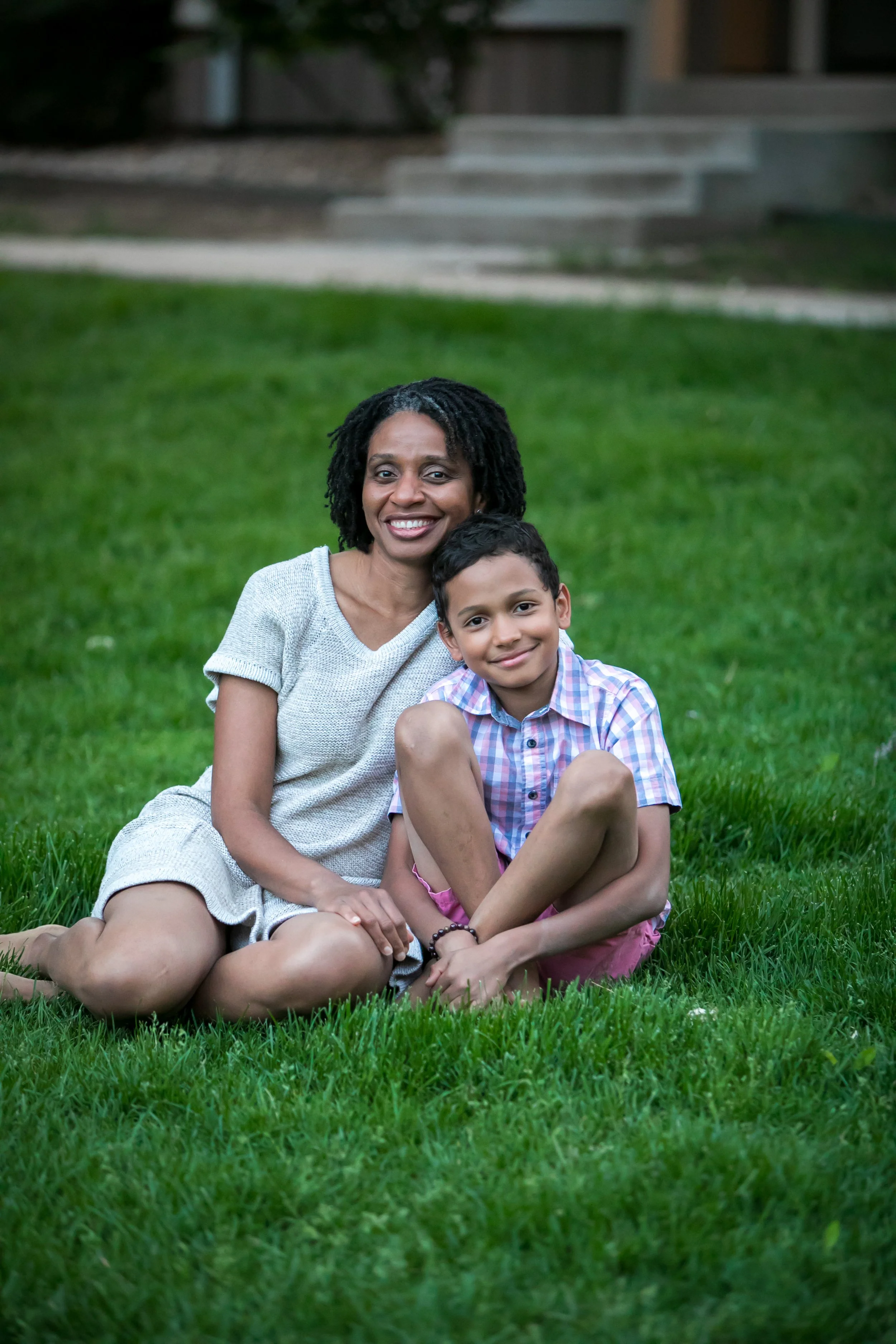 A woman and a boy sitting on green grass, smiling at the camera.