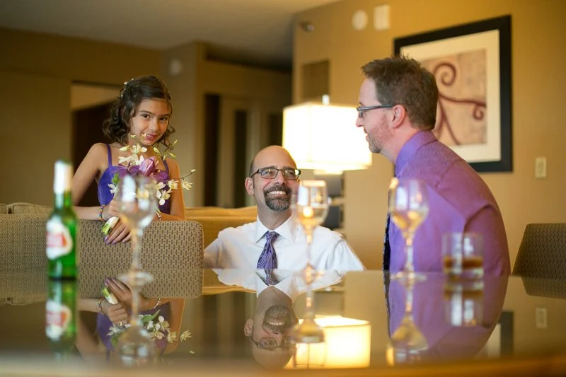 A girl giving a flower bouquet to a smiling man in a white shirt, sitting at a table with two glasses of wine, a beer, and a bottle, in a warmly lit room with artwork on the wall.