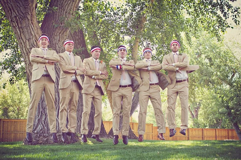 Group of six men in beige suits wearing red, white, and blue headbands, standing with arms crossed in a park with trees and a wooden fence in the background.