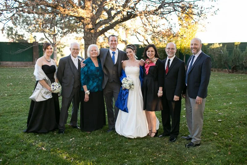 Family celebrating at a wedding outdoors on a lawn during sunset, with a tree in the background. The bride is in a white gown holding a bouquet, and the groom is in a dark suit with a blue tie. Six family members are around them, dressed in formal at