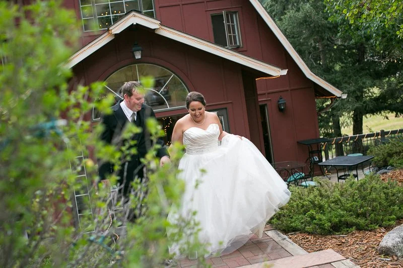 A bride in a white wedding dress and a groom in a suit happily walking outside a red barn with greenery around.
