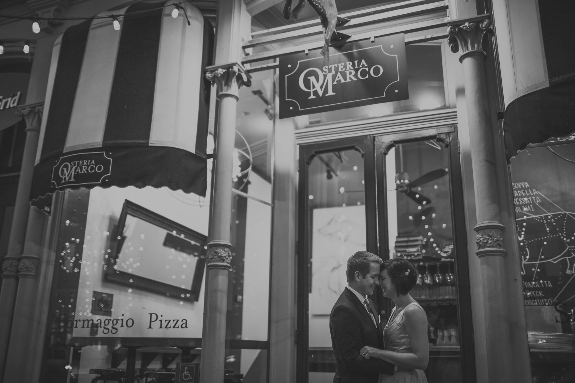A black and white photo of a couple close together, smiling and touching foreheads, standing outside a restaurant called Osteria Marco. The restaurant sign is visible, and the couple appears happy and intimate.
