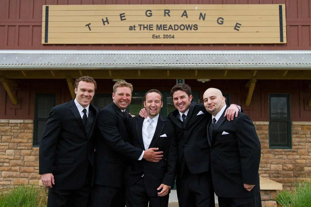 Five men dressed in suits standing outside a building with a sign that reads 'The Grange at The Meadows, Est. 2004'. They are smiling and have their arms around each other.