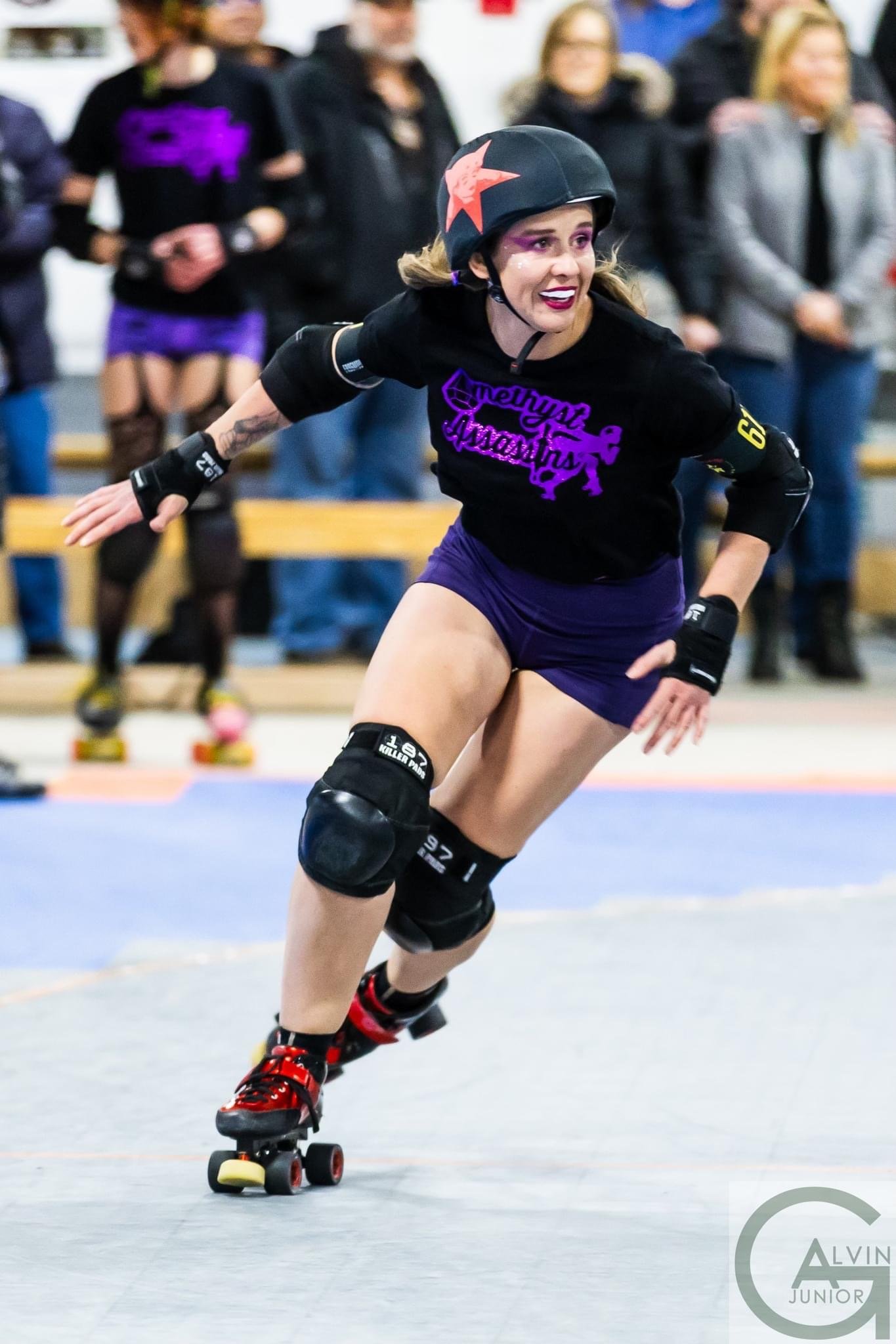 A woman wearing roller derby gear, including a helmet and knee pads, skating on an indoor track with spectators in the background.