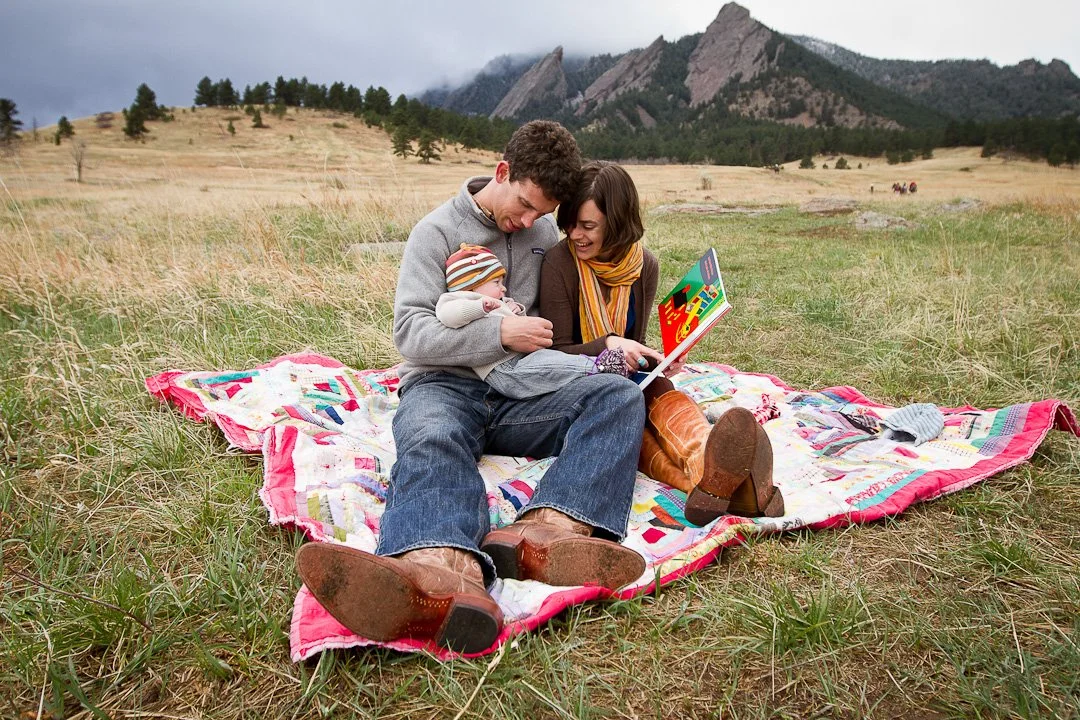 A family with a baby sitting on a blanket in an open field with mountains in the background, reading a book together.