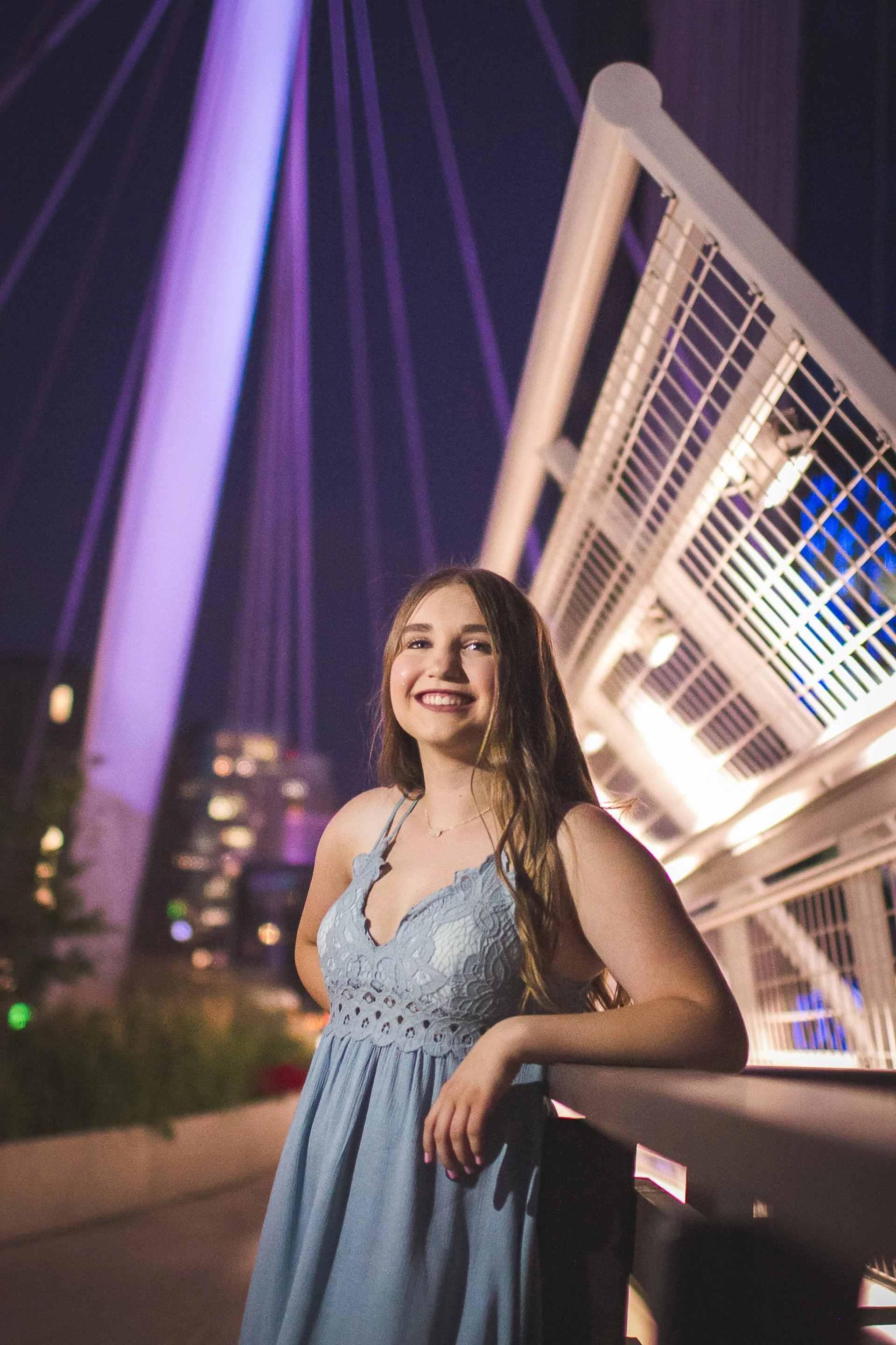 A young woman smiling at night, standing outdoors with a modern illuminated bridge and city skyline in the background.