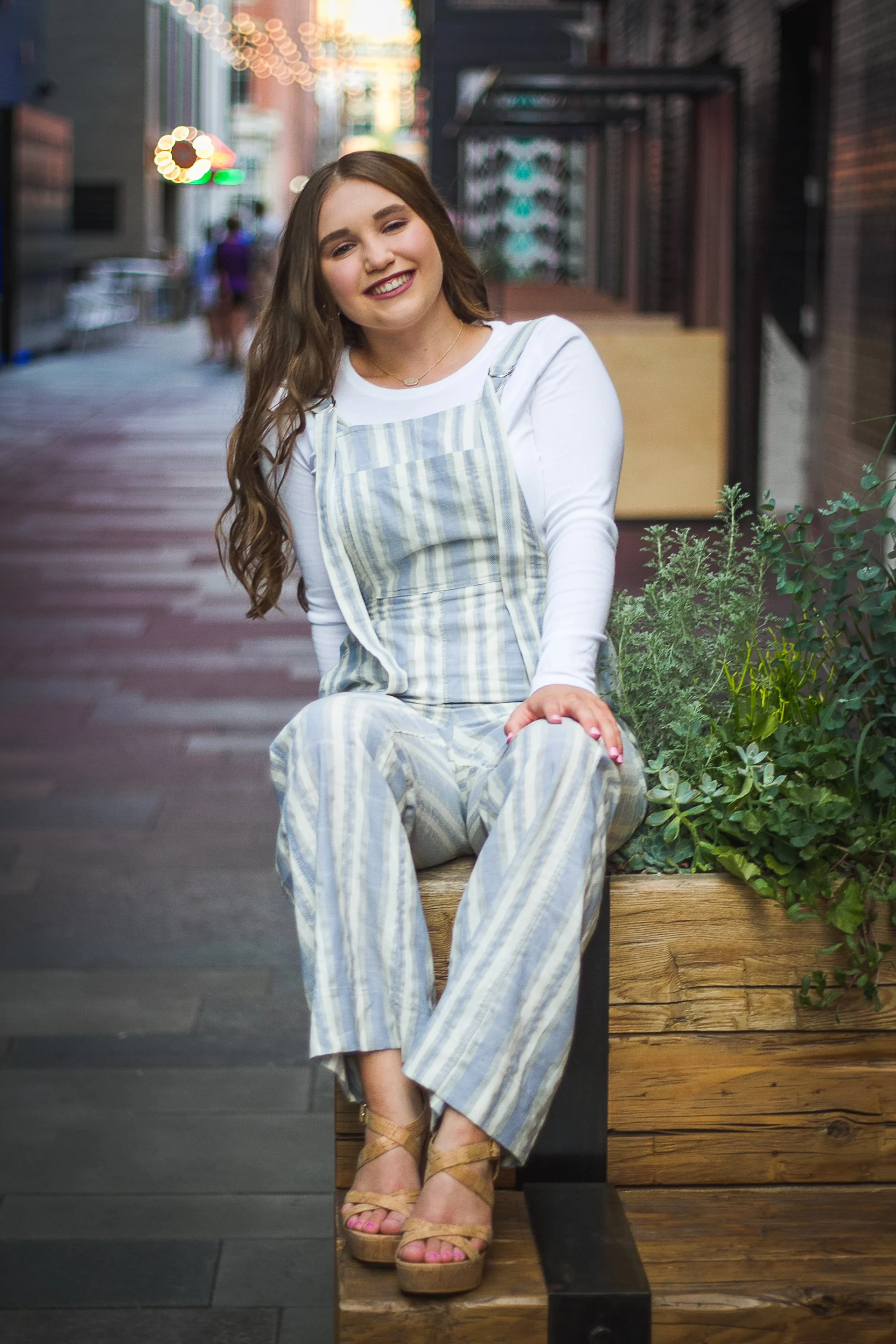 A young woman with long brown hair sitting on a wooden bench on a city sidewalk, smiling and wearing a striped outfit and tan wedge sandals.