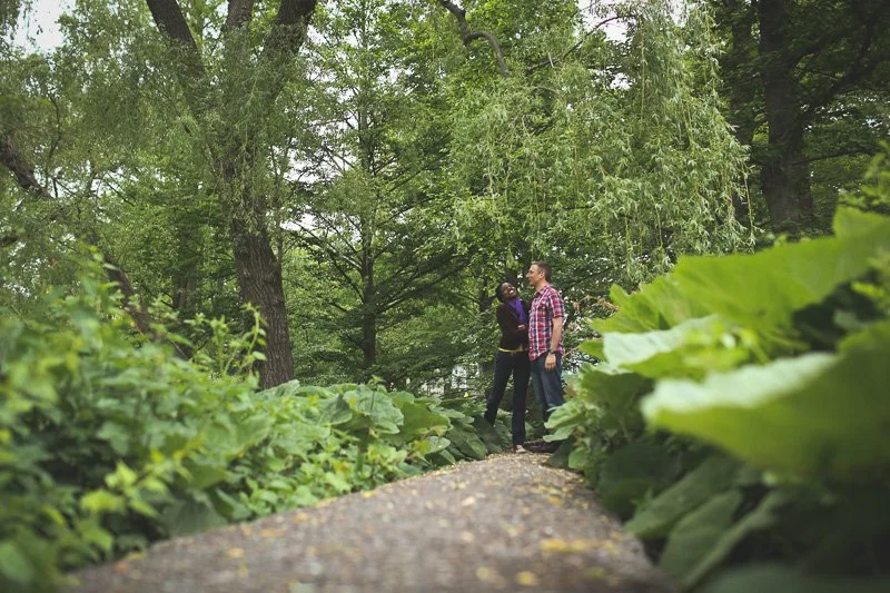 A man and woman standing and talking on a wooded trail surrounded by green trees and plants.