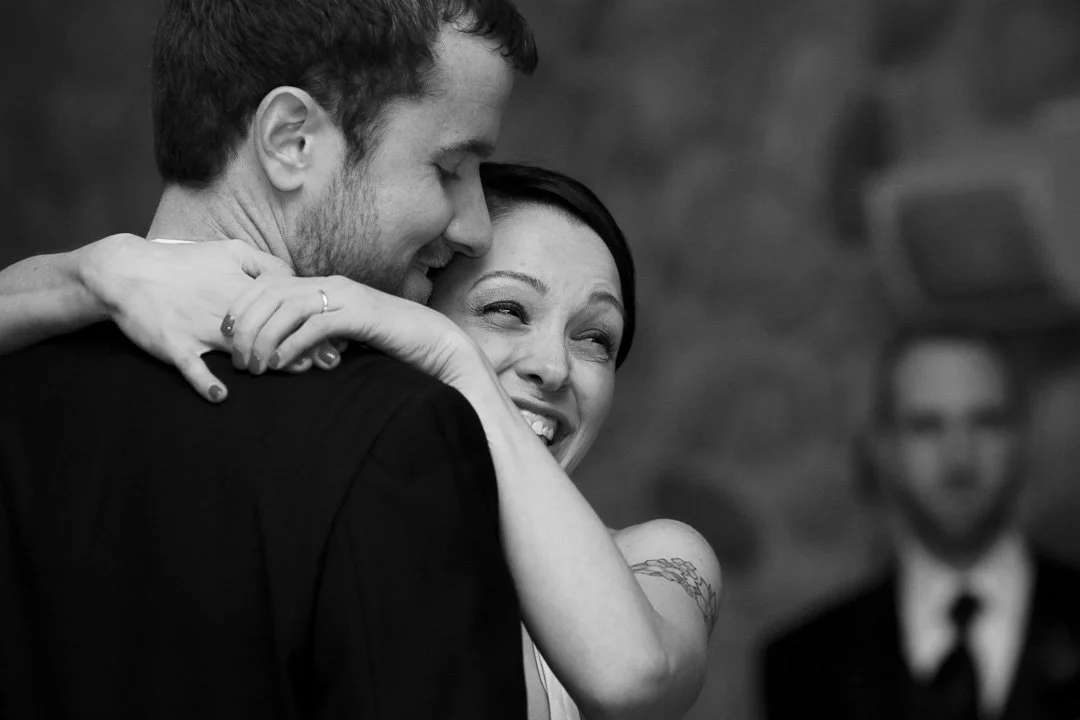 A black and white photo of a joyful couple hugging, with a man with dark hair and beard and a woman with dark hair smiling and hugging him tightly. In the background, there is a man with glasses and a beard in a suit, slightly out of focus.
