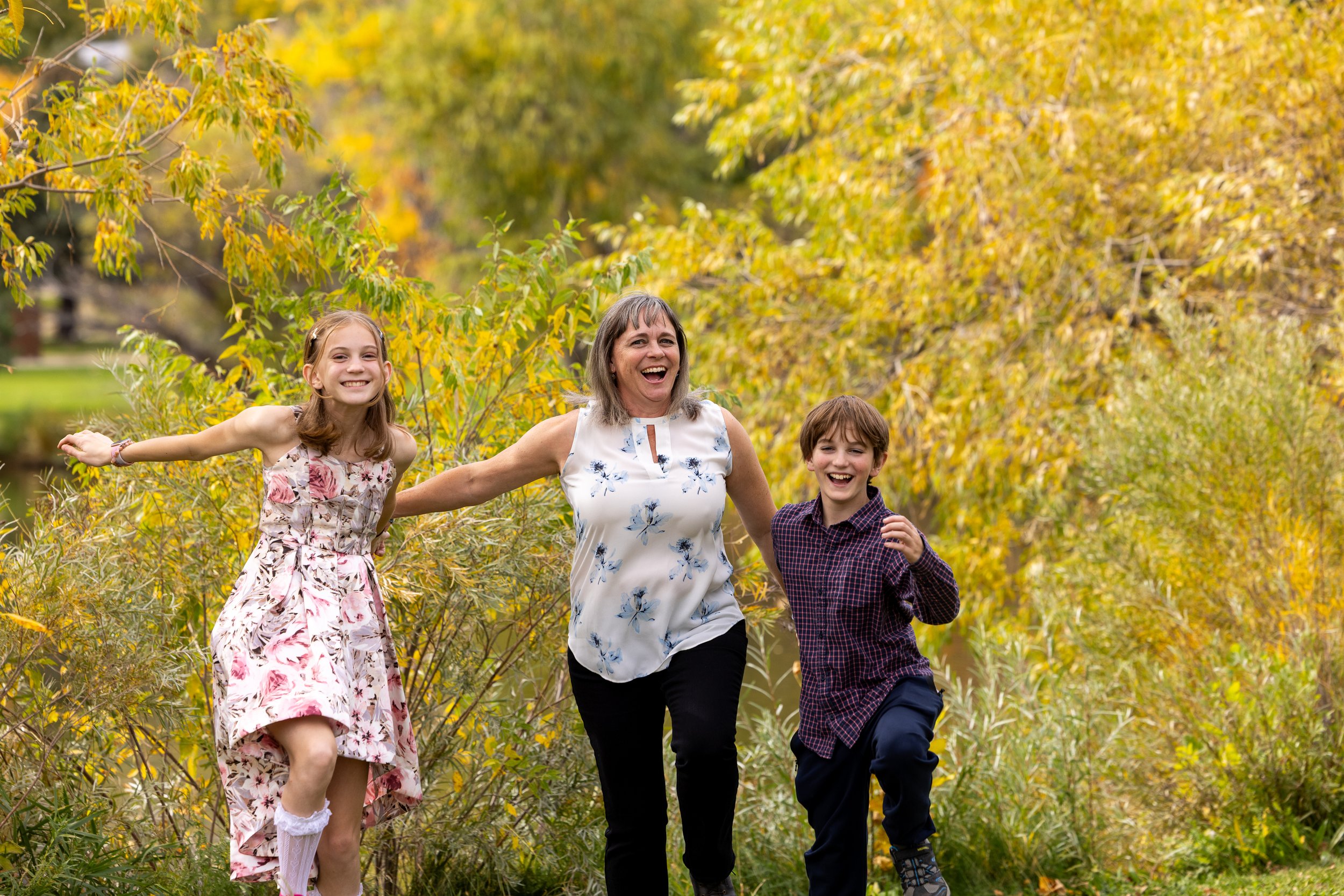 A woman and two children, a girl and a boy, are running and smiling through a park with yellow and green autumn foliage.