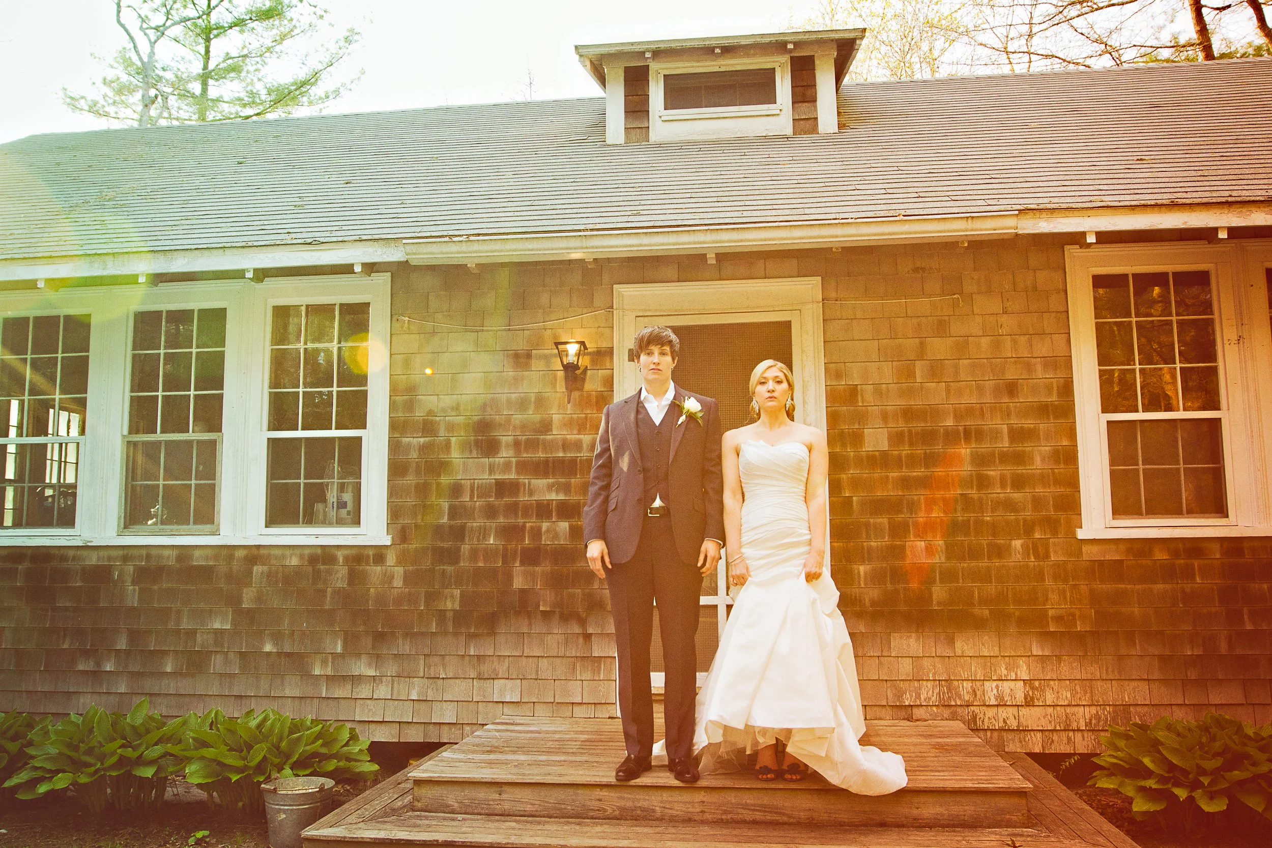 A bride and groom stand side by side on a small wooden porch in front of a house, lit by warm sunlight.