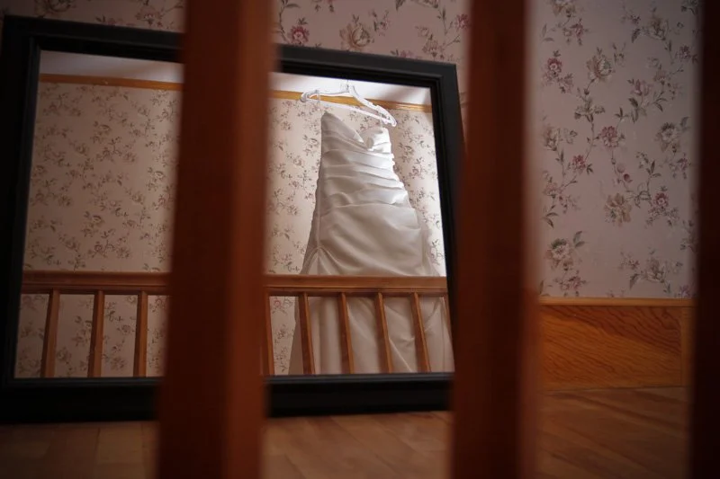 A wedding dress hanging on a hanger inside a black frame, viewed through the wooden slats of a crib or bed, with wallpapered wall in the background.