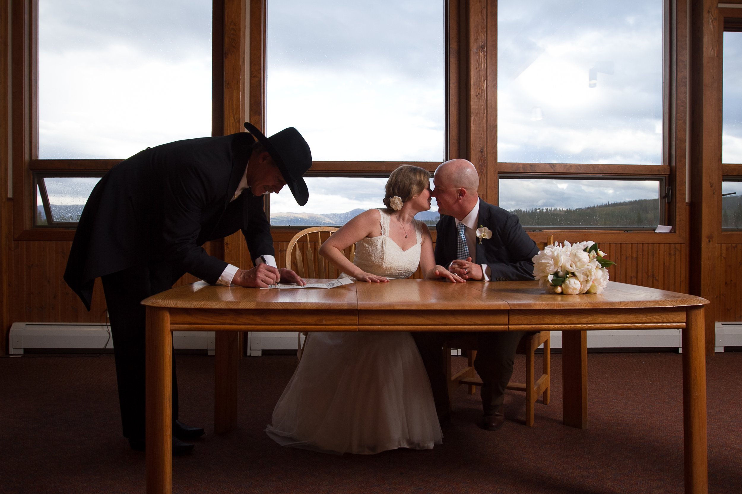 Couple getting married seated at a wooden table, with an officiant signing a document, and exchanging vows, in a rustic indoor setting with large windows showing a cloudy mountain landscape.