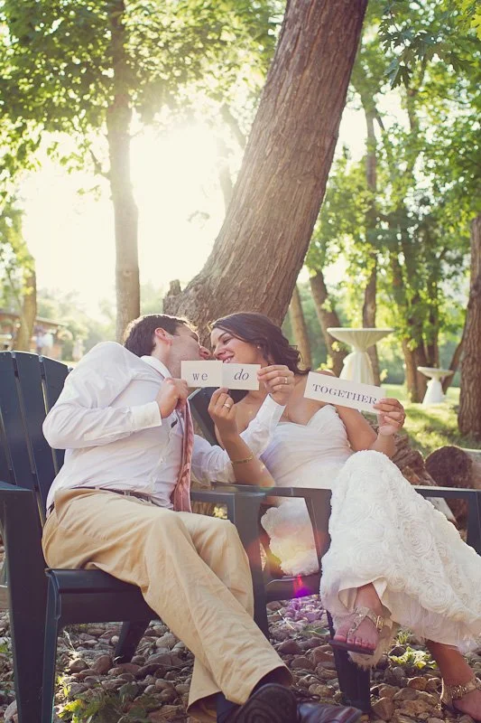 A couple is sitting outdoors on black chairs, sharing a romantic moment. They are holding a sign that says 'We do,' with the woman wearing a white dress and the man in a white shirt and tan pants. Sunlight filters through trees in a lush green park s