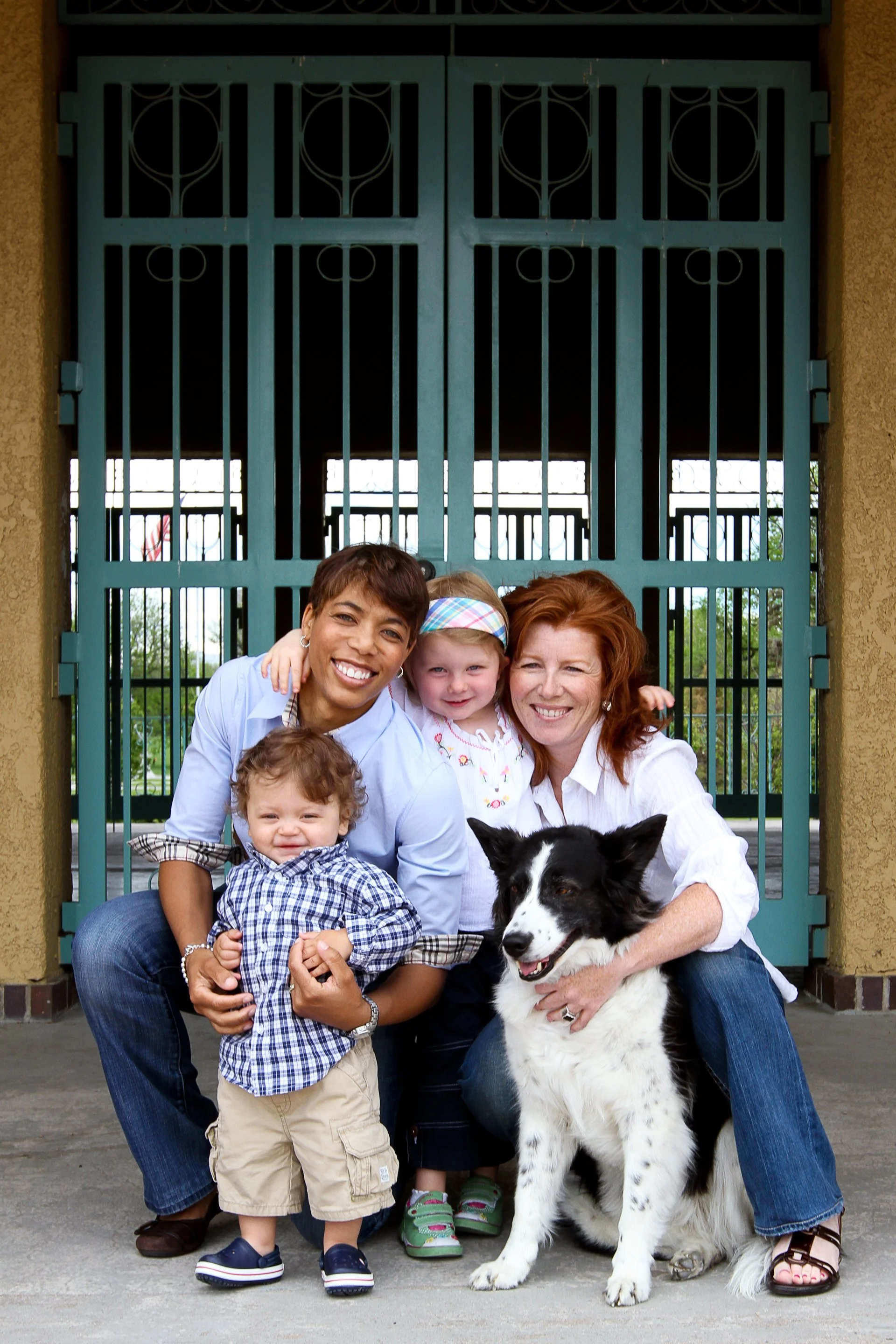A group of people, including two women and two children, along with a black and white dog, smiling and posing in front of a teal gate.