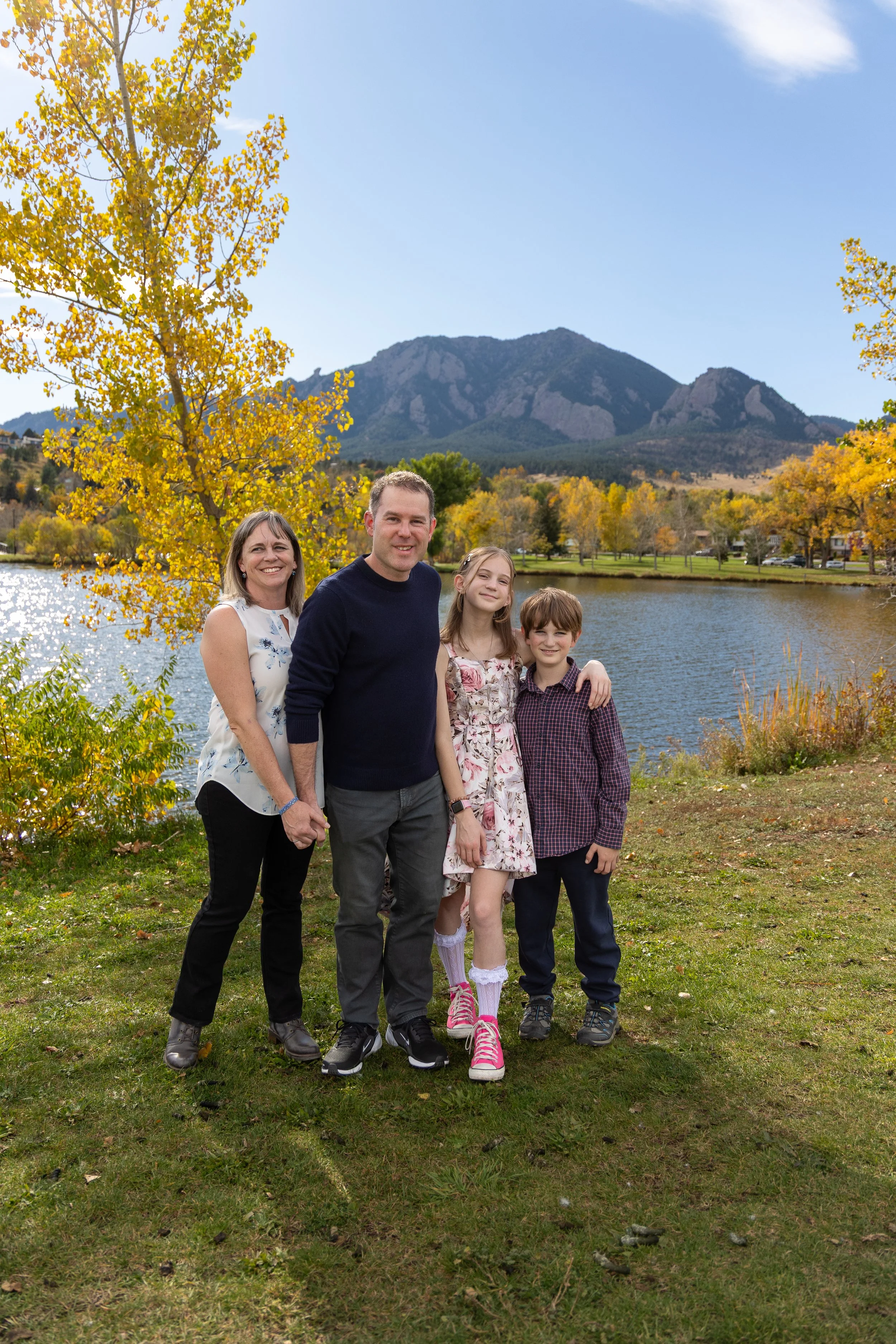 Family of four standing together outdoors near a lake with mountains in the background during daytime in autumn, with trees showing fall colors.