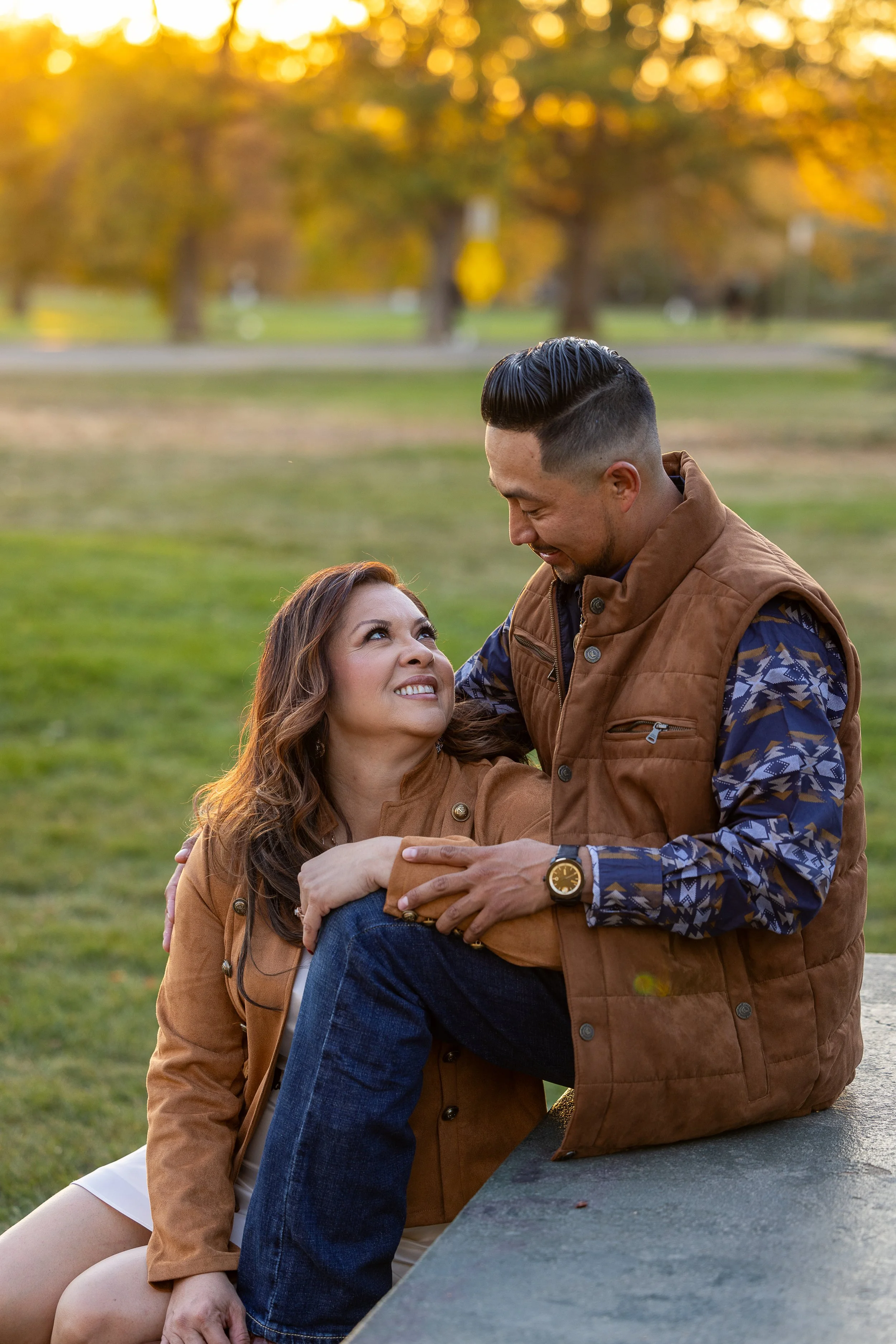 A woman sitting on the grass and a man sitting on a concrete surface in an outdoor park during sunset, looking at each other lovingly.