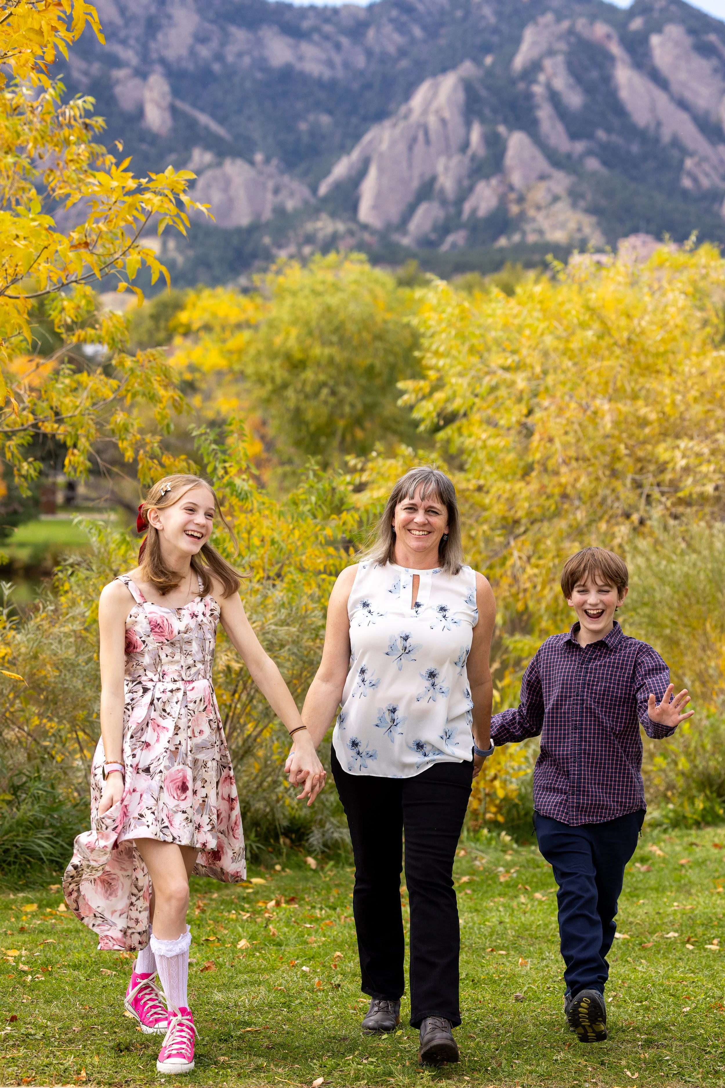 A woman with two children, a girl and a boy, holding hands and walking on a grassy path with autumn trees and mountains in the background. All are smiling and enjoying the outdoor fall setting.