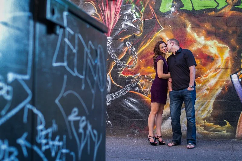 A couple standing close together in front of a colorful graffiti mural with a skull and fiery background, with a graffiti-covered wall in the foreground.