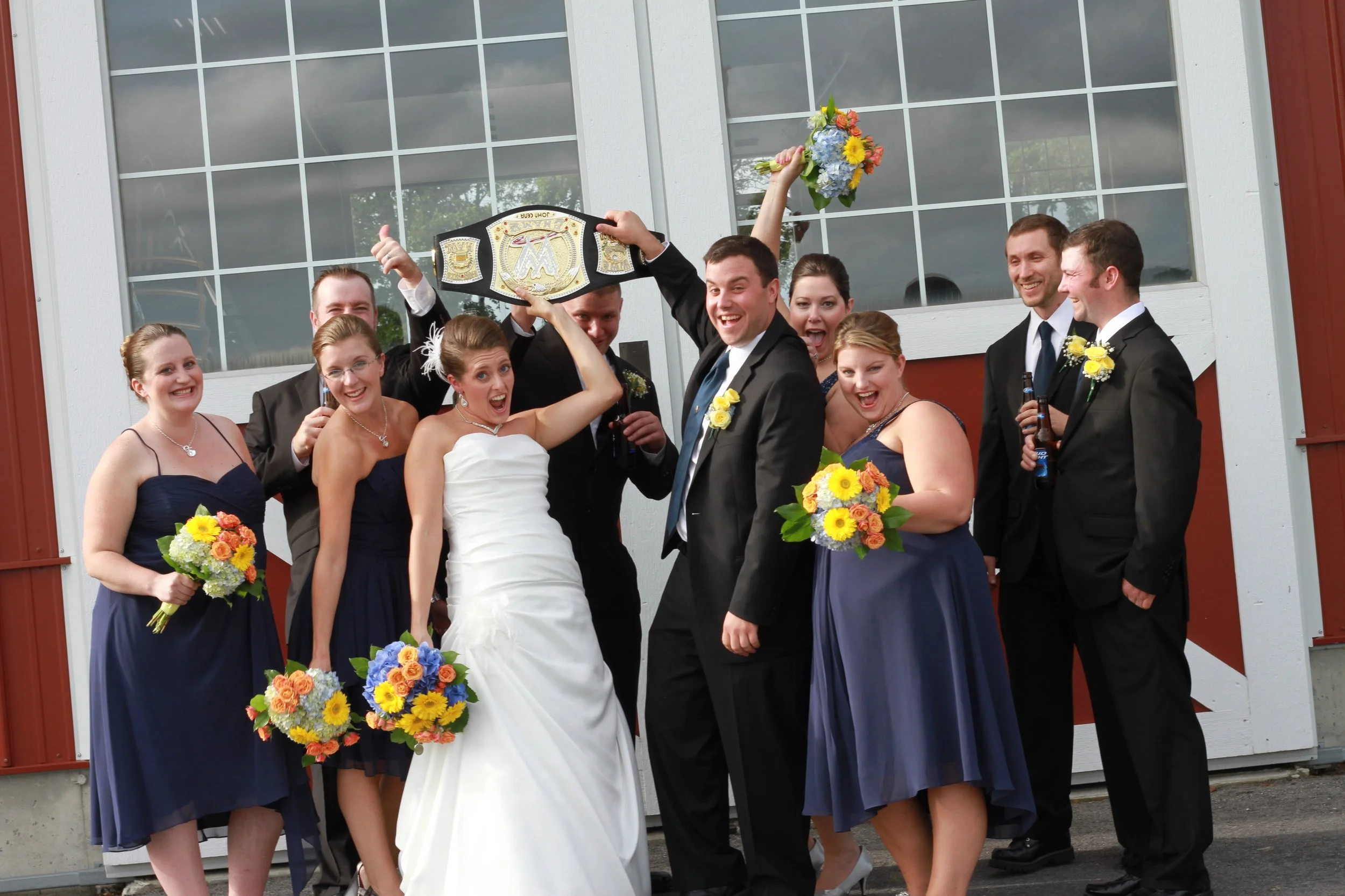 Group of wedding guests celebrating outside, holding bouquets, with some in formal attire, and a man holding a championship belt.