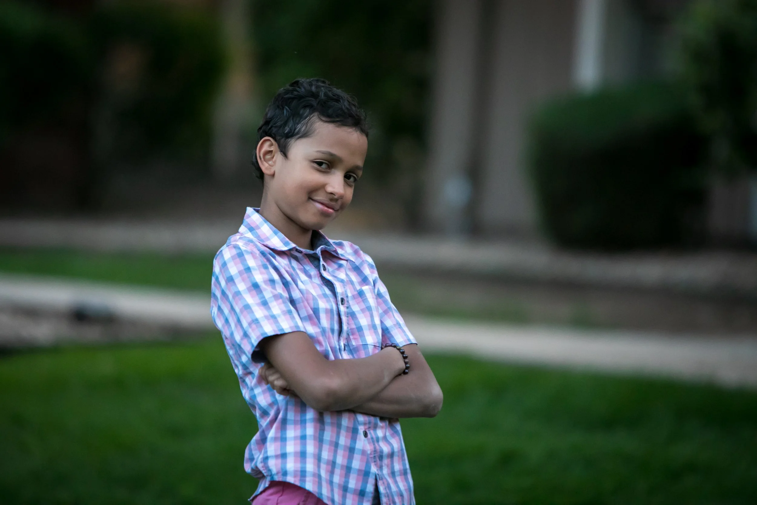 A young boy with dark, curly hair stands outdoors on a grassy area, arms crossed, wearing a colorful checkered shirt and a black bracelet, with a slight smile.